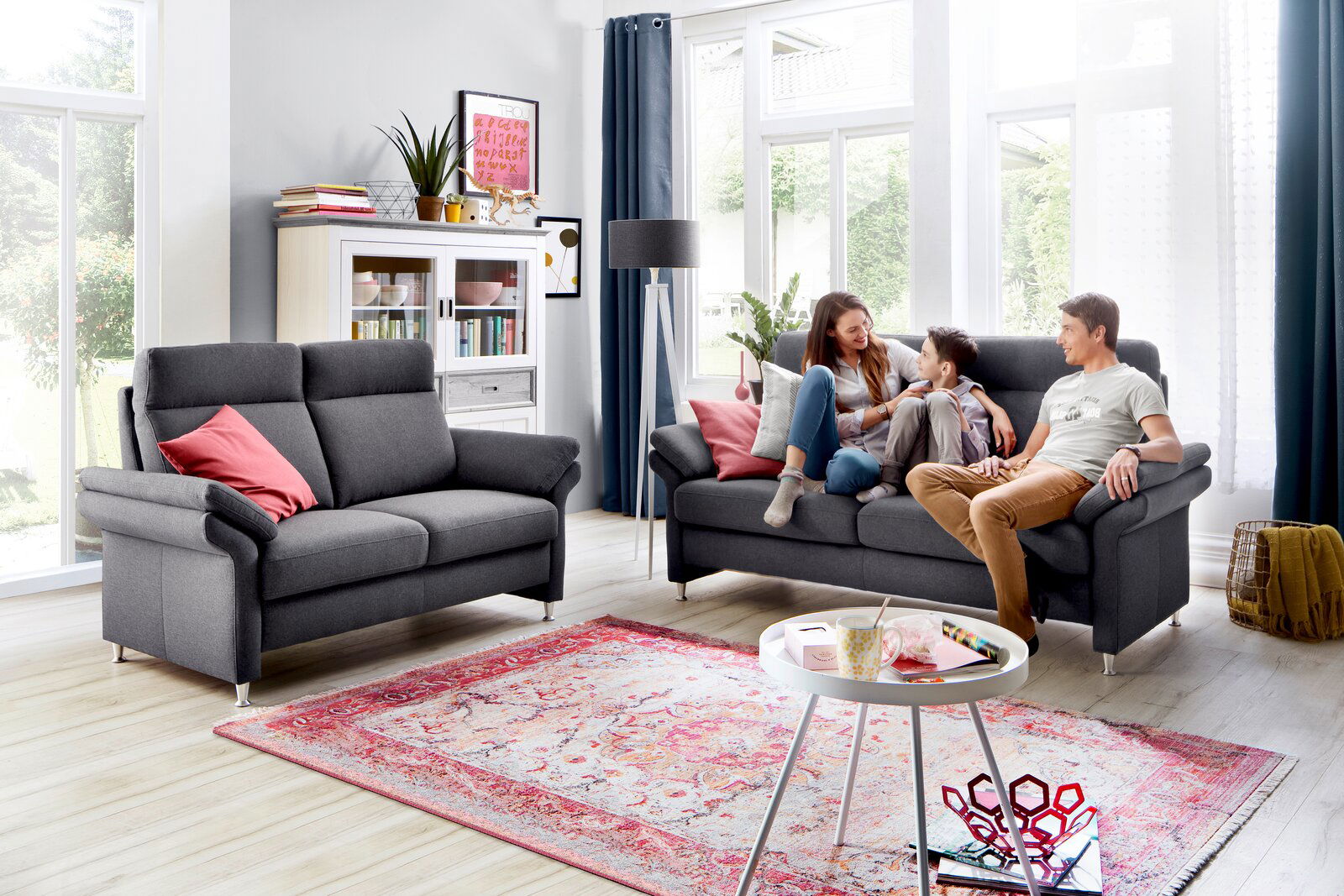 Wohnzimmer mit zwei grauen Sofas, eines davon mit einer Familie darauf, im Hintergrund ein weißes Regal und eine Stehlampe, Perspektive von vorne.