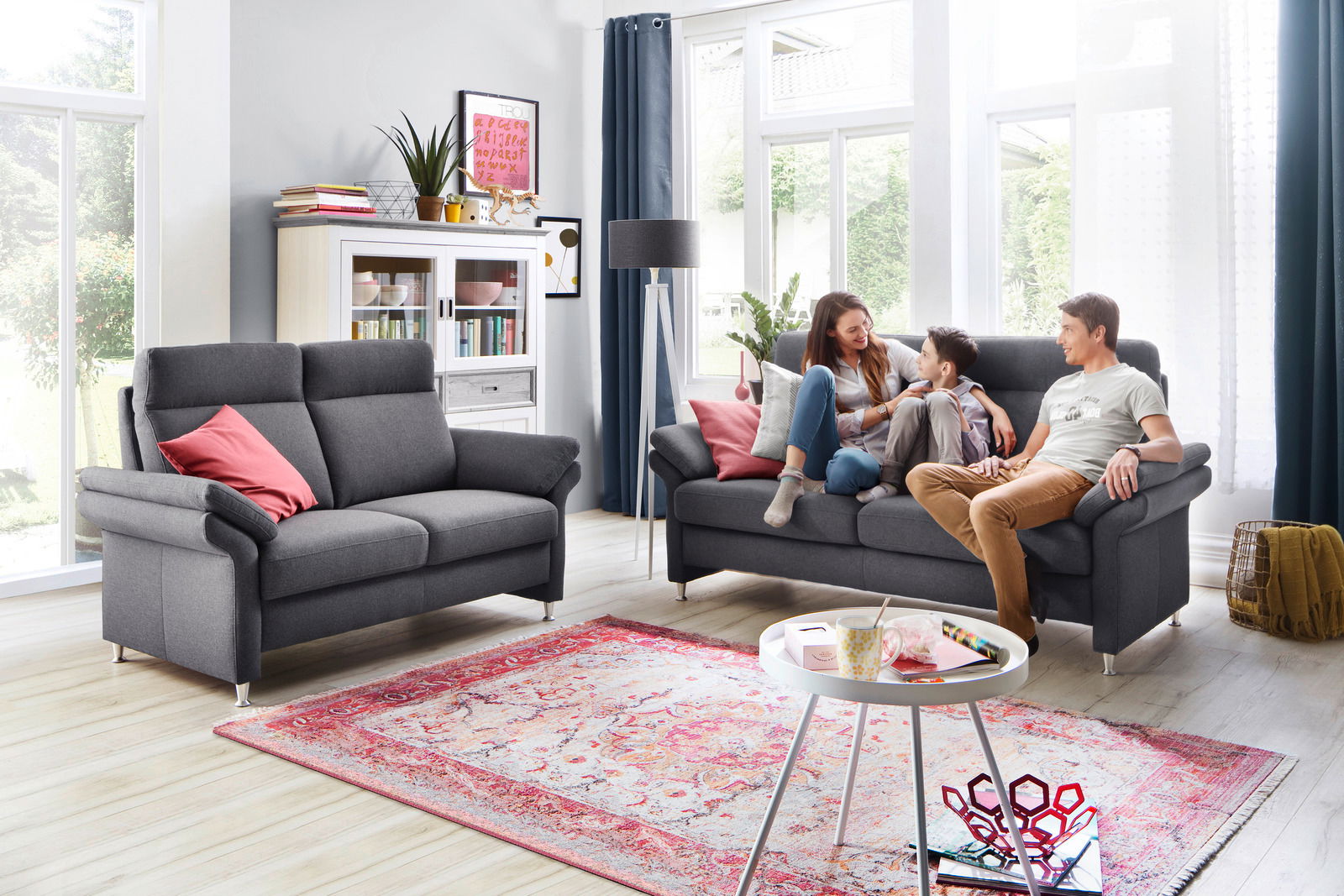 Wohnzimmer mit zwei grauen Sofas, eines davon mit einer Familie darauf, im Hintergrund ein weißes Regal und eine Stehlampe, Perspektive von vorne.