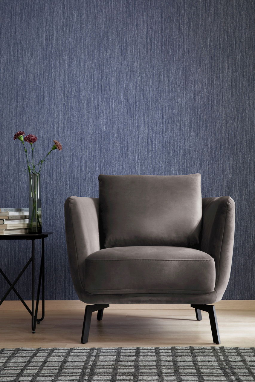 Grauer Sessel in Frontalansicht vor einer blauen Wand, neben einem Beistelltisch mit Büchern und Blumen.