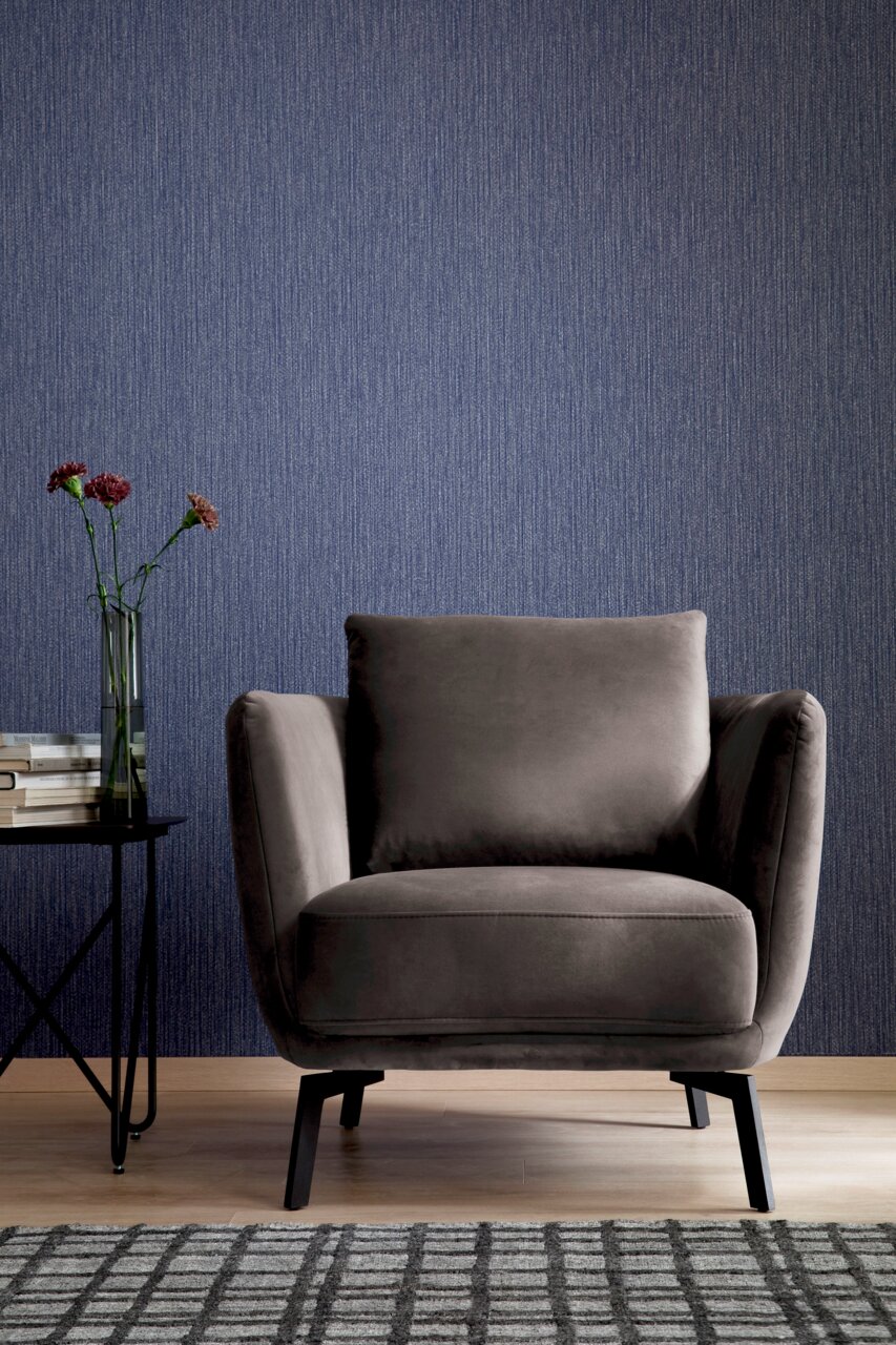 Grauer Sessel in Frontalansicht vor einer blauen Wand, neben einem Beistelltisch mit Büchern und Blumen.