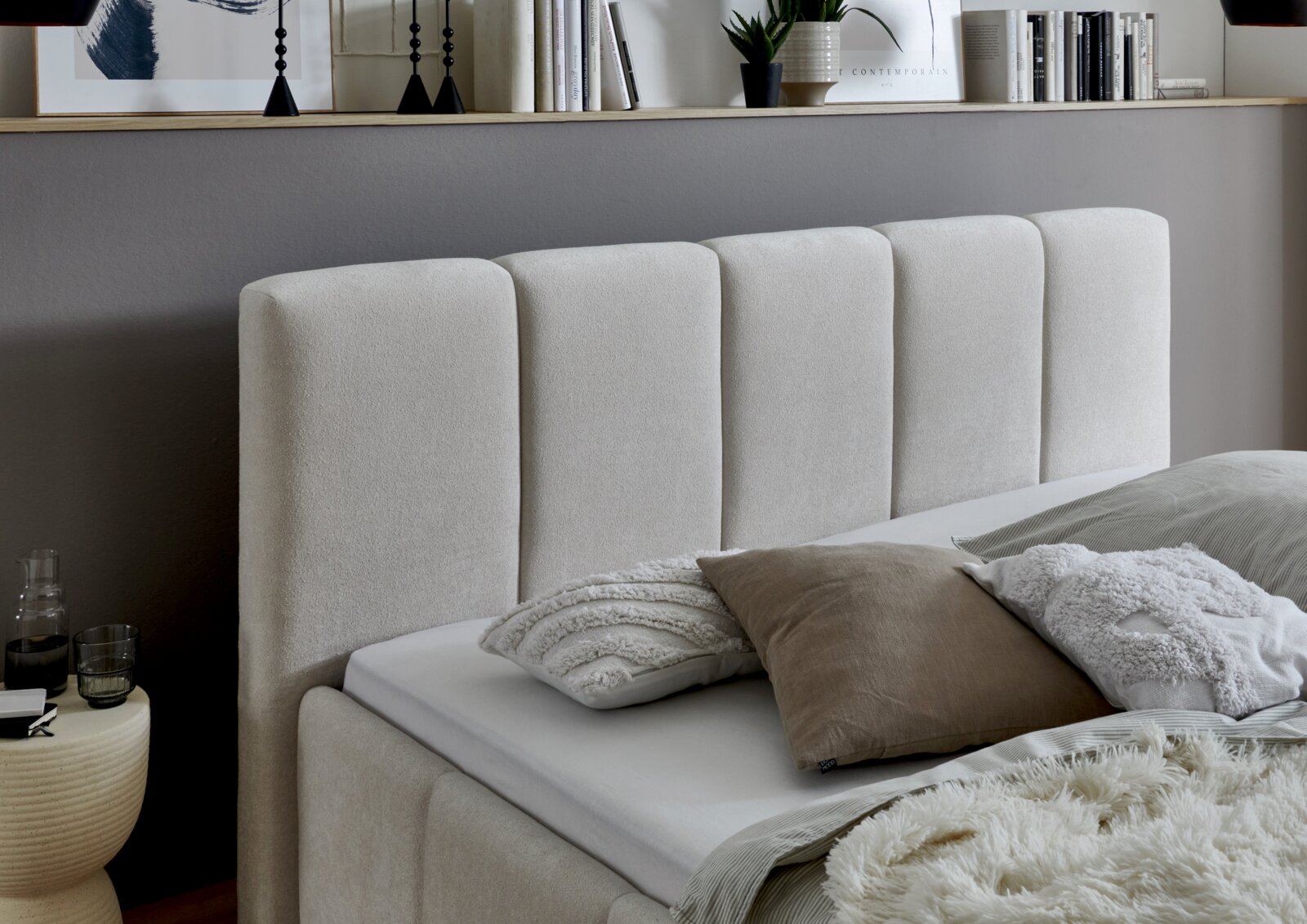 Zijaanzicht van een modern gestoffeerd bed met een beige hoofdeinde en decoratieve kussens.