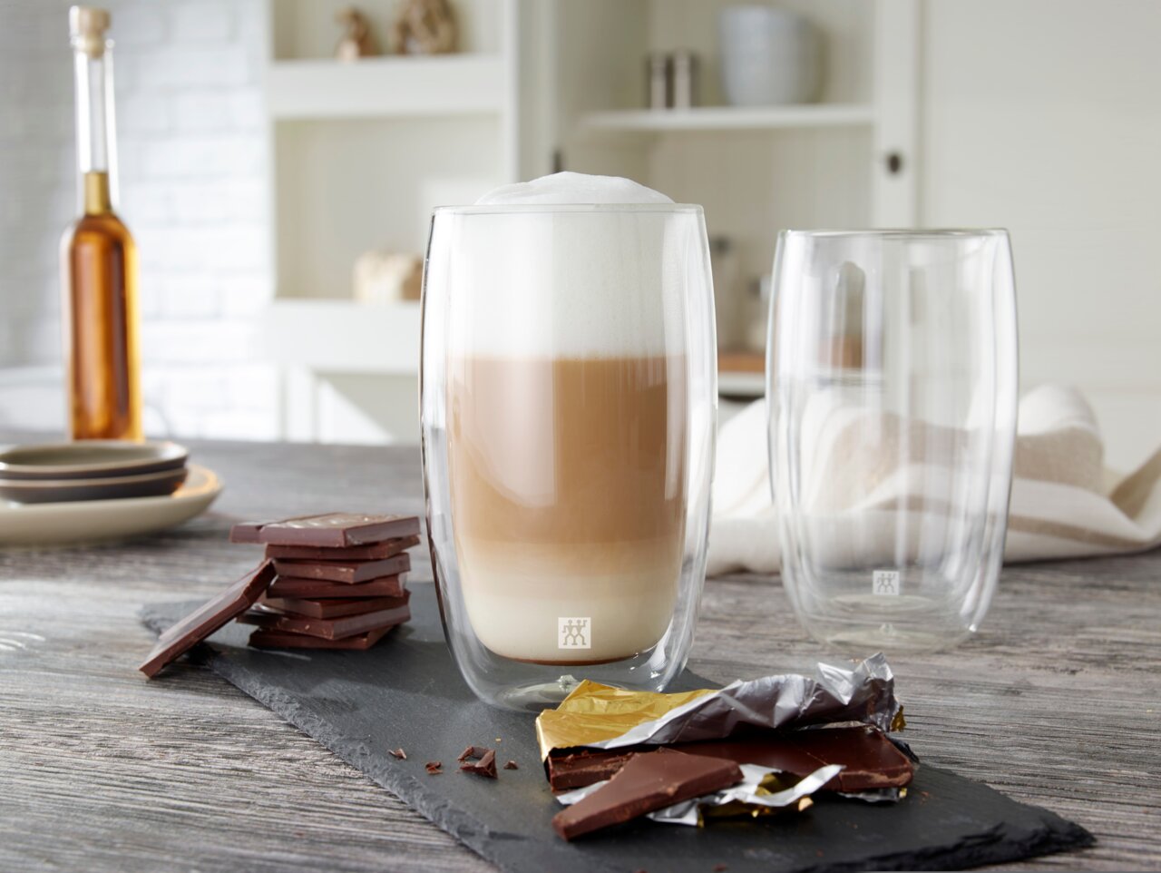 Twee dubbelwandige latte macchiato-glazen, waarvan één gevuld met een gelaagde latte macchiato, op een tafel met stukjes chocolade, gezien vanuit een zijaanzicht.