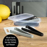 Verschlussclips in verschiedenen Farben auf einem Tisch, mit einer transparenten Aufbewahrungsbox im Hintergrund. Perspektive von oben.