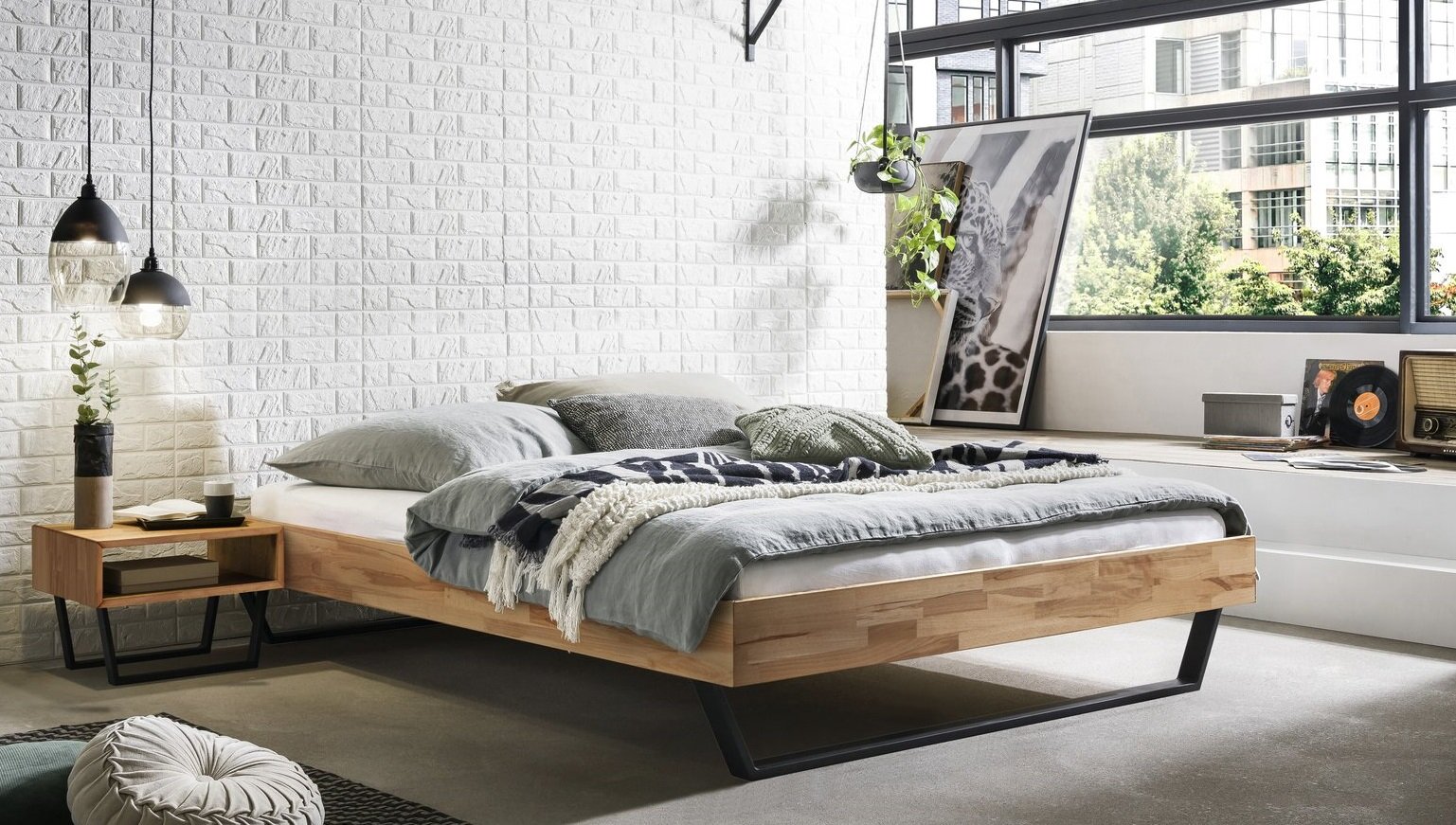 Modernes Holz-Bettgestell im Industrial-Stil mit schwarzem Metallkufenfuß, bezogen mit grauer Bettwäsche in einem hellen Loft-Schlafzimmer Perspektive: seitliche Vorderansicht leicht von links.