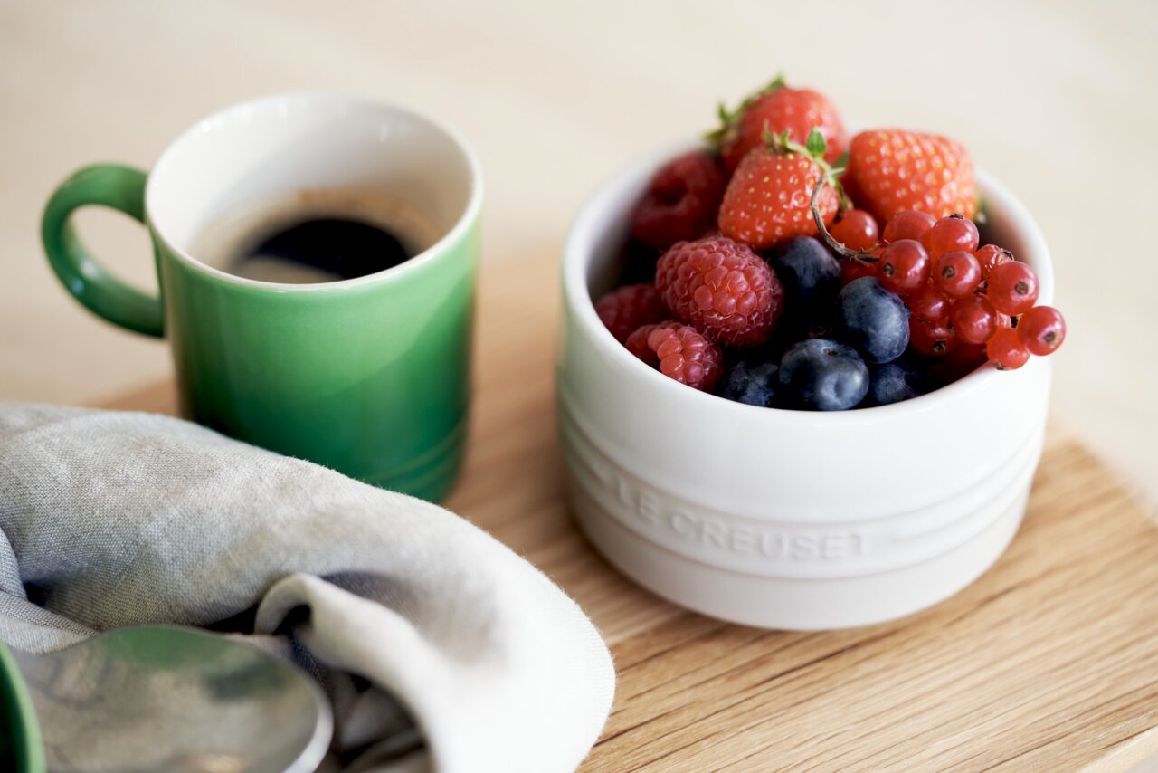 Grüner Becher mit Kaffee neben einer weißen Schale voller frischer Beeren auf einem Holztisch, von oben betrachtet.