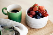 Grüner Becher mit Kaffee neben einer weißen Schale voller frischer Beeren auf einem Holztisch, von oben betrachtet.