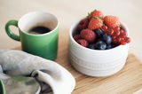Grüner Becher mit Kaffee neben einer weißen Schale voller frischer Beeren auf einem Holztisch, von oben betrachtet.