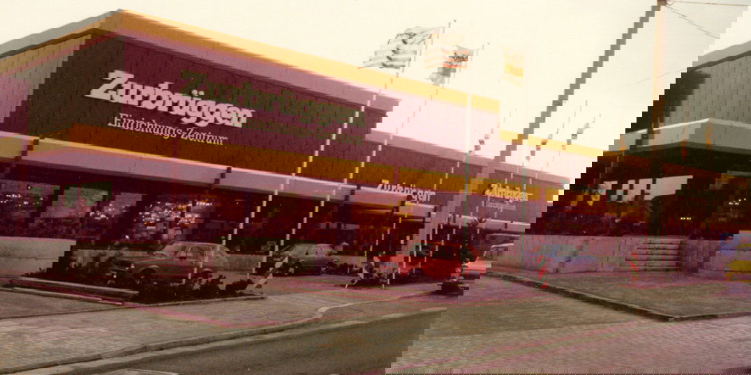 Zurbrüggen Verkaufshaus in Holzwickede. Vor dem Gebäude ist ein kleiner Parkplatz. Ein rotes und ein blaues Auto parkt dort.