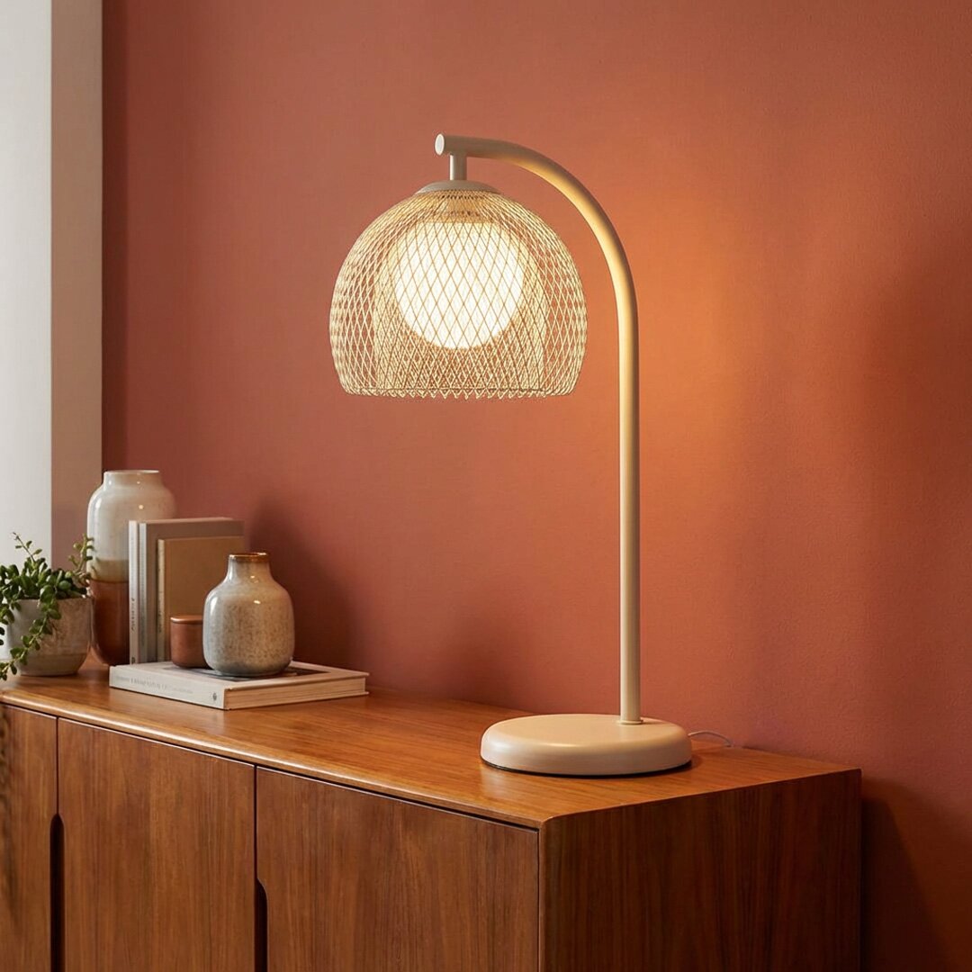 Schöner Wohnen Tafellamp MESH Beige Tischlampe mit gebogenem Standfuß und halbrundem, geflochtenem Schirm, eingeschaltet auf Holzsideboard vor terrakottafarbener Wand, Frontansicht leicht von links.