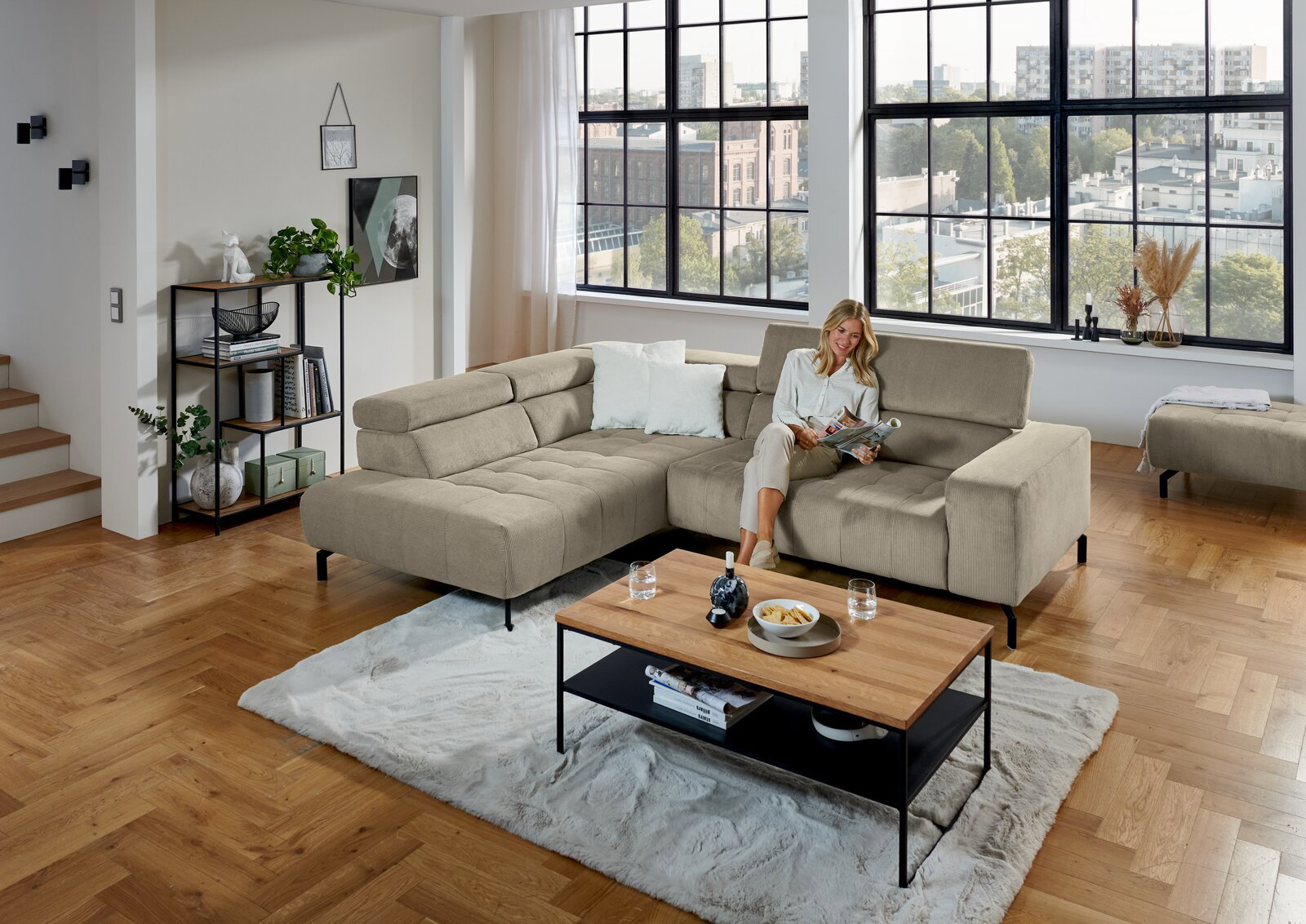 Beiges L-förmiges Ecksofa mit breiter Ottomane und schwarzen Metallfüßen in modernem Loft-Wohnzimmer, frontale leicht erhöhte Perspektive; Frau sitzt entspannt mit Magazin, davor Couchtisch auf hellem Teppich.