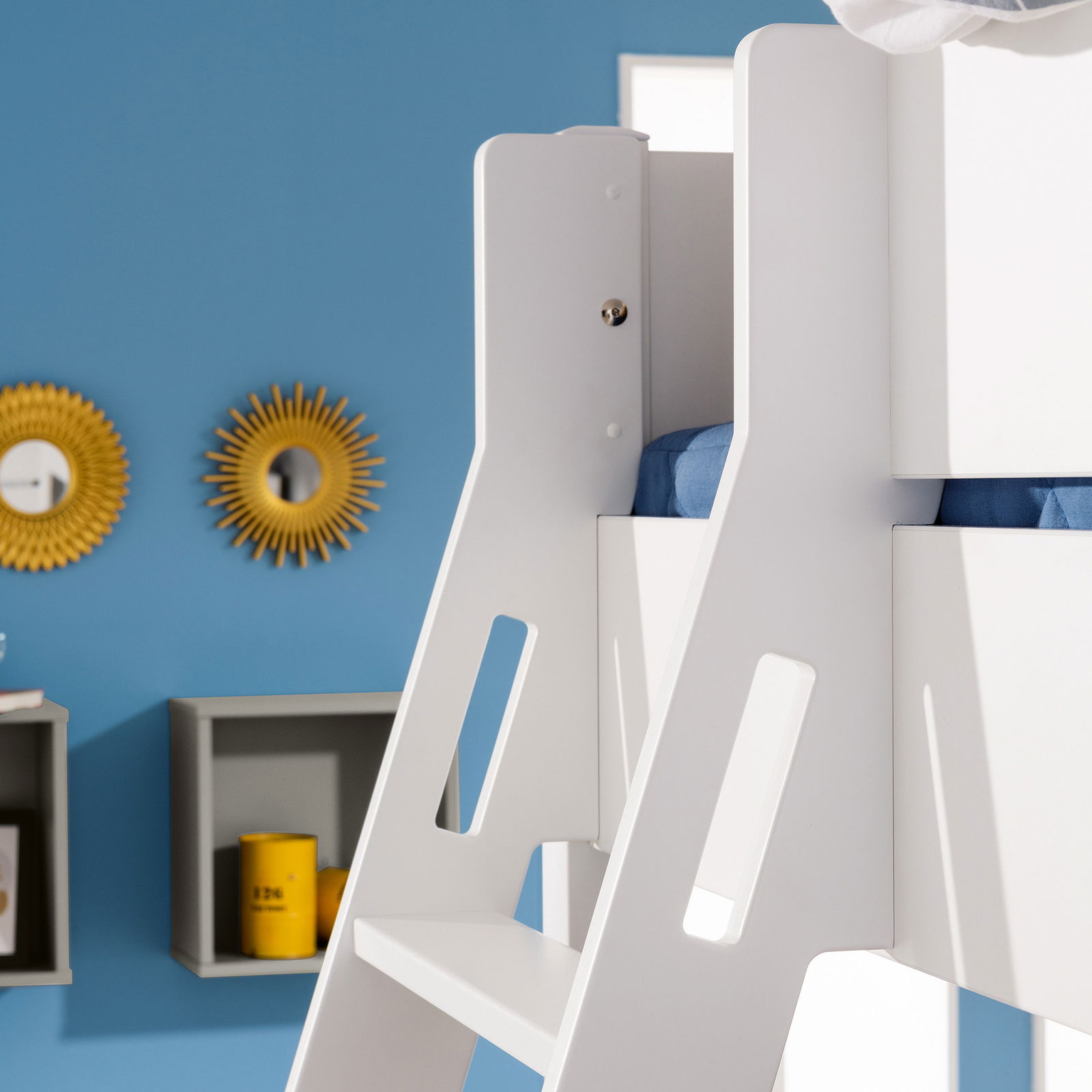 Nahaufnahme eines weißen Spielbetts mit blauer Bettwäsche, seitliche Perspektive, im Hintergrund blaue Wand mit dekorativen Spiegeln.