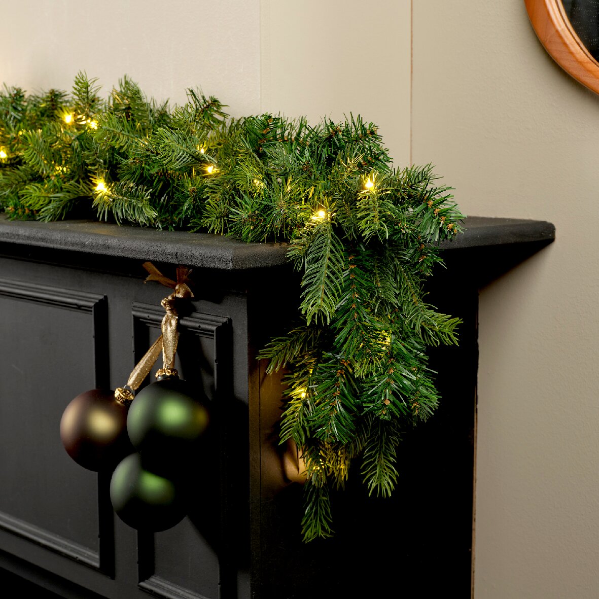 Schuin vooraanzicht van een weelderige groene kerstguirlande met warmwitte verlichting, gedrapeerd over een zwarte schouw en versierd met donkergroene en bruine kerstballen
