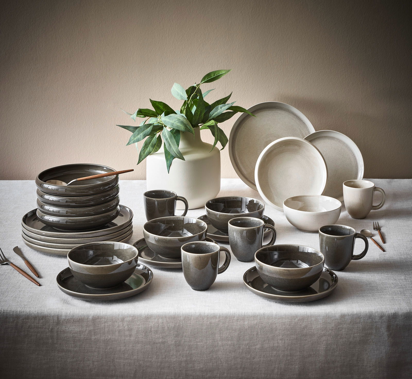 Kombiservice 20-tlg. CANJU Moonbeamblomus, bestehend aus Tellern, Tassen und Schalen in neutralen Tönen, auf einem Tisch mit einer Pflanze im Hintergrund, aus seitlicher Perspektive fotografiert.