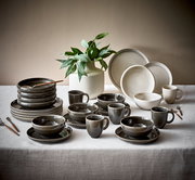 Kombiservice 20-tlg. CANJU Moonbeamblomus, bestehend aus Tellern, Tassen und Schalen in neutralen Tönen, auf einem Tisch mit einer Pflanze im Hintergrund, aus seitlicher Perspektive fotografiert.