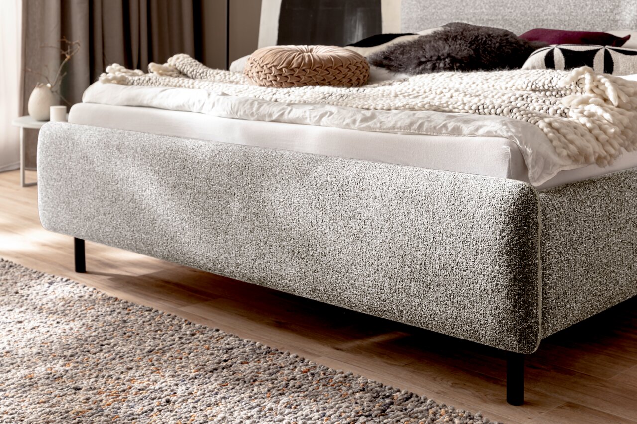 Zijaanzicht van een grijs gestoffeerd bed met sierkussens en dekens op een houten vloer.