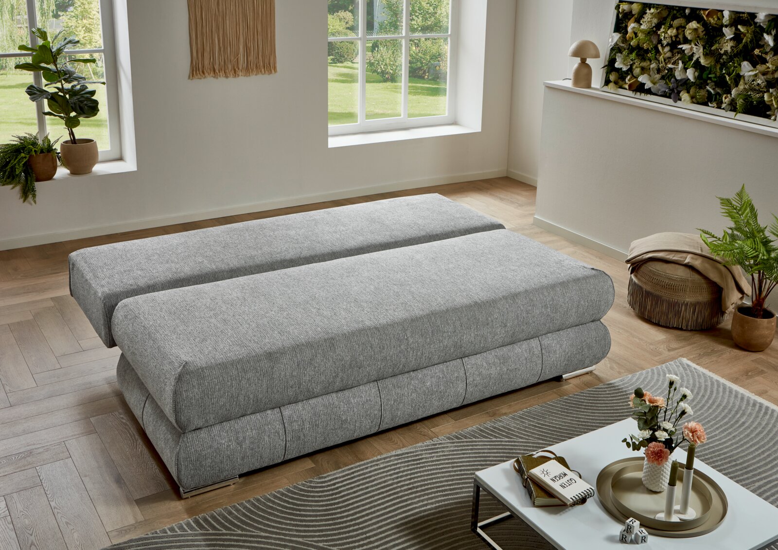 Modernes graues Schlafsofa/Polsterbett in einem hellen Wohnzimmer, Perspektive: leicht erhöhte schräge Seitenansicht von vorne.