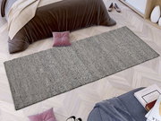 Handgewebter Teppich in Grau, rechteckig, auf hellem Holzboden vor einem Bett mit brauner Decke, aus der Vogelperspektive.