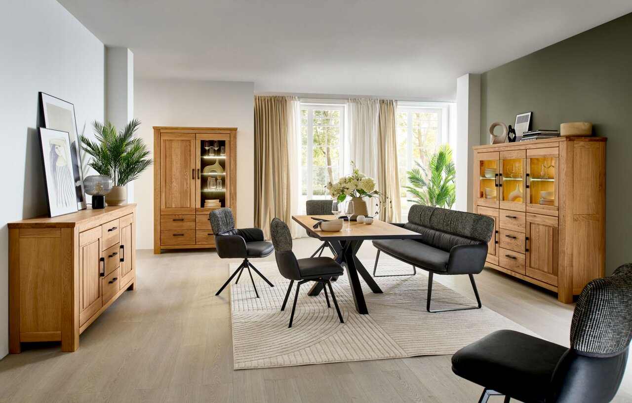 Moderne eetkamer met massief eikenhouten meubels (dressoir, vitrinekast en hoge kast met glazen deuren en verlichting), houten eettafel met zwarte stoelen en banken op een lichtgekleurd tapijt, gefotografeerd vanuit een breedhoekperspectief.