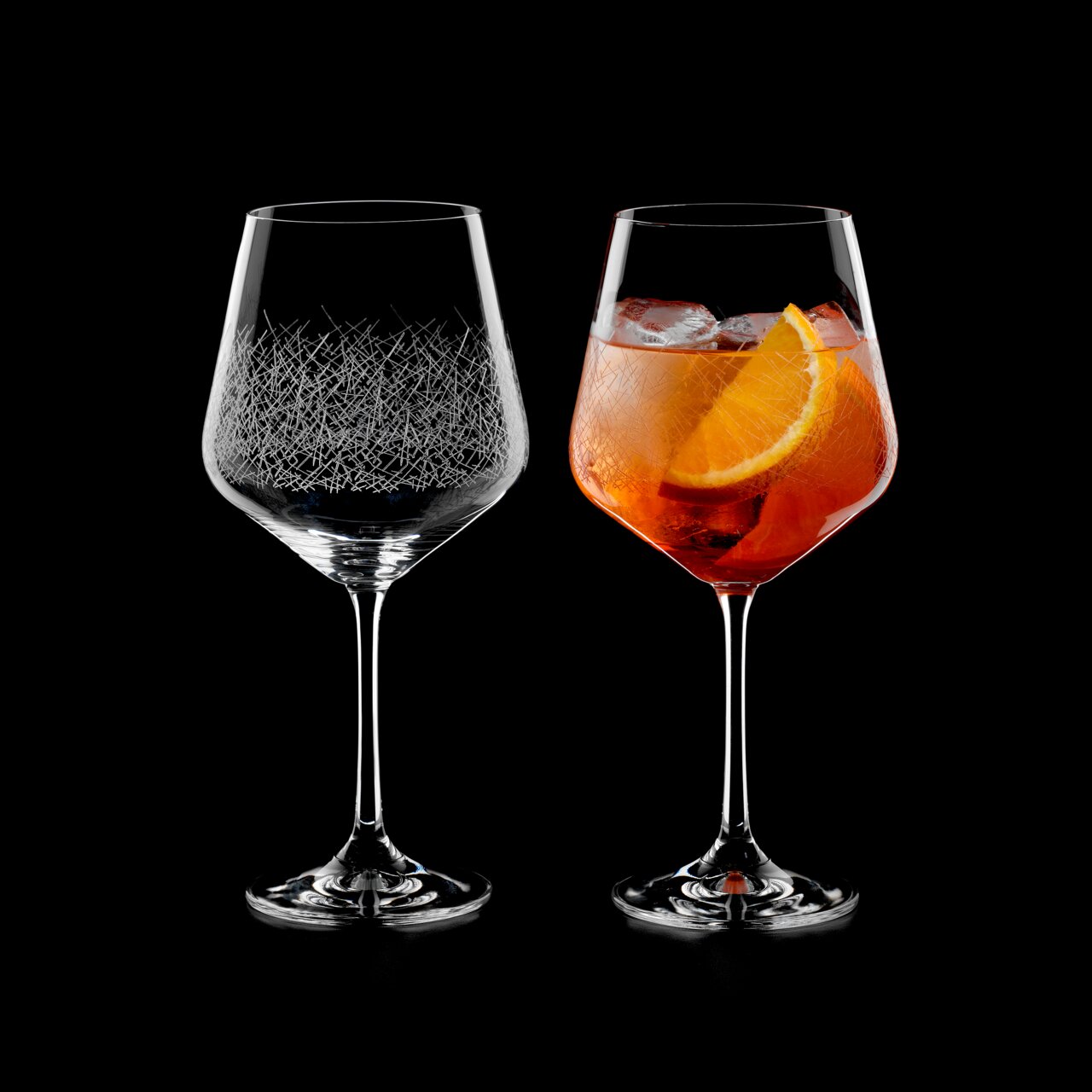 Twee elegante cocktailglazen, één leeg en één gevuld met een drankje en een schijfje sinaasappel, op een zwarte achtergrond, gezien vanuit een zijdelingse hoek.