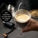 Nahaufnahme eines doppelwandigen Thermo-Cappuccino-Glases, das in der Hand gehalten wird, mit dampfendem Kaffee und einer Kaffeekanne im Hintergrund.