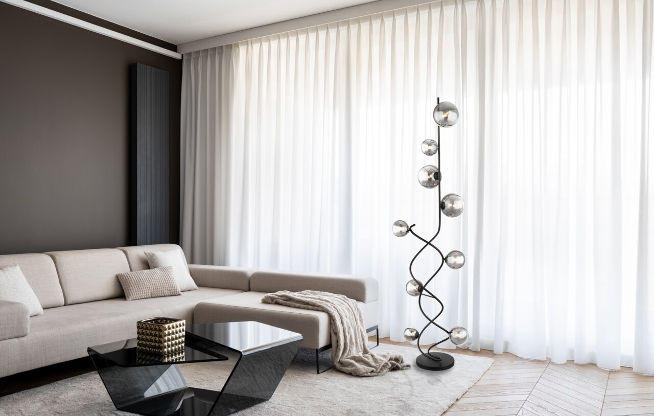 Moderne Stehleuchte mit mehreren kugelförmigen Leuchten auf einem geschwungenen schwarzen Gestell, im Wohnzimmer neben einem beigen Sofa und vor weißen Vorhängen, seitliche Perspektive.