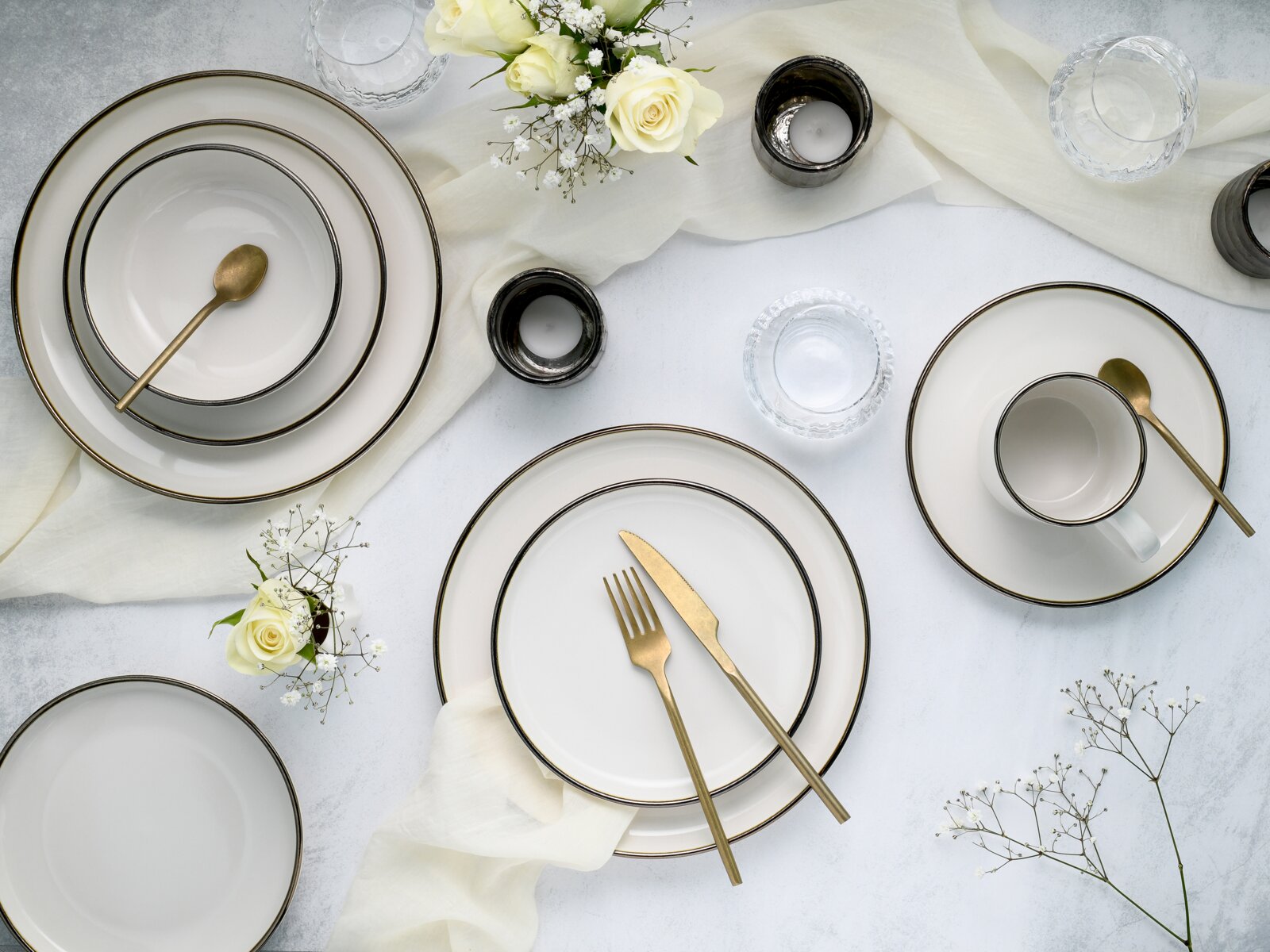 Draufsicht auf elegant gedeckten Tisch mit weißem Geschirr mit goldfarbenem Rand, goldfarbenem Besteck, Teetasse, Wassergläsern, Kerzenhaltern und weißen Rosen auf hellem Untergrund