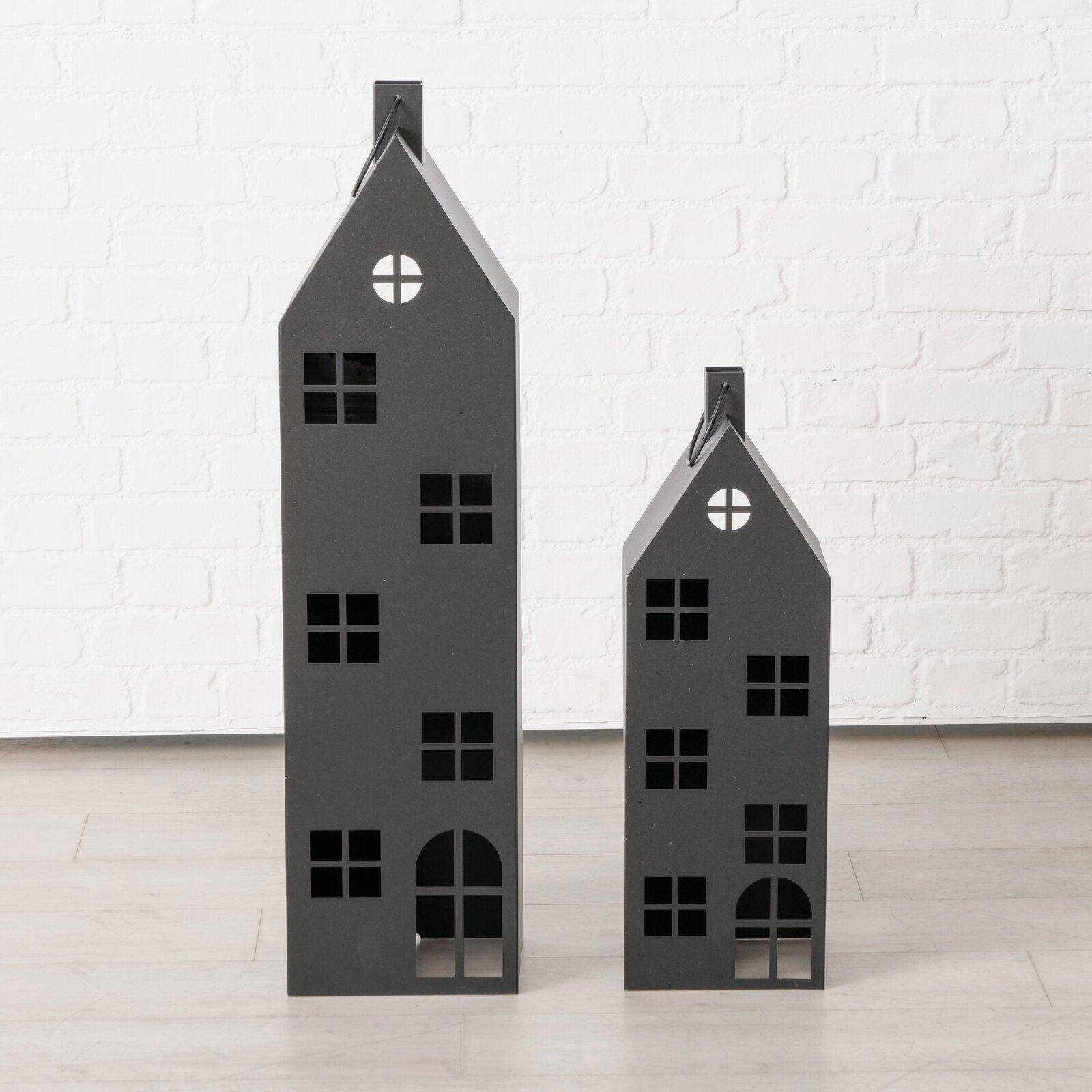 Twee zwarte, huisvormige metalen lantaarns van verschillende grootte, met raamopeningen en een handvat, gefotografeerd van voren op een lichte achtergrond tegen een witte bakstenen muur.
