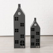 Twee zwarte, huisvormige metalen lantaarns van verschillende grootte, met raamopeningen en een handvat, gefotografeerd van voren op een lichte achtergrond tegen een witte bakstenen muur.