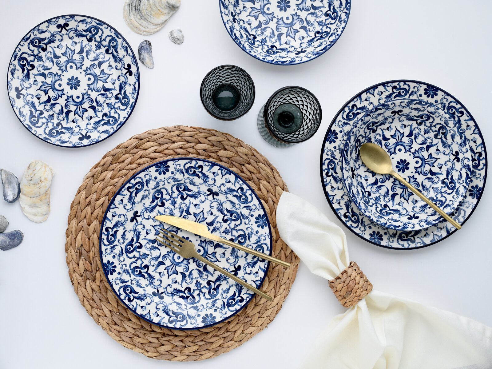 Draufsicht (Flatlay) auf blau-weiß gemustertes Porzellan-Geschirrset mit Tellern und Schalen, ergänzt durch goldfarbenes Besteck, zwei dunkle Trinkgläser, geflochtene Platzsets und Serviette auf hellem Hintergrund.