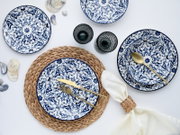 Draufsicht (Flatlay) auf blau-weiß gemustertes Porzellan-Geschirrset mit Tellern und Schalen, ergänzt durch goldfarbenes Besteck, zwei dunkle Trinkgläser, geflochtene Platzsets und Serviette auf hellem Hintergrund.