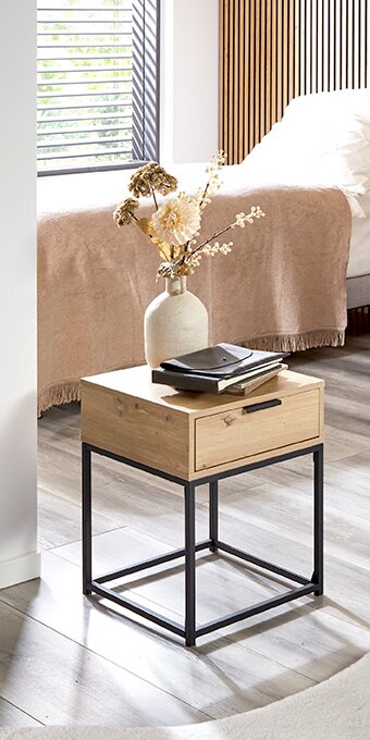 Moderner Nachttisch/Beistelltisch aus hellem Holz mit schwarzem Metallgestell und Schublade, dekoriert mit Vase und Büchern, in einem hellen Schlafzimmer – Perspektive: leicht schräg von vorne