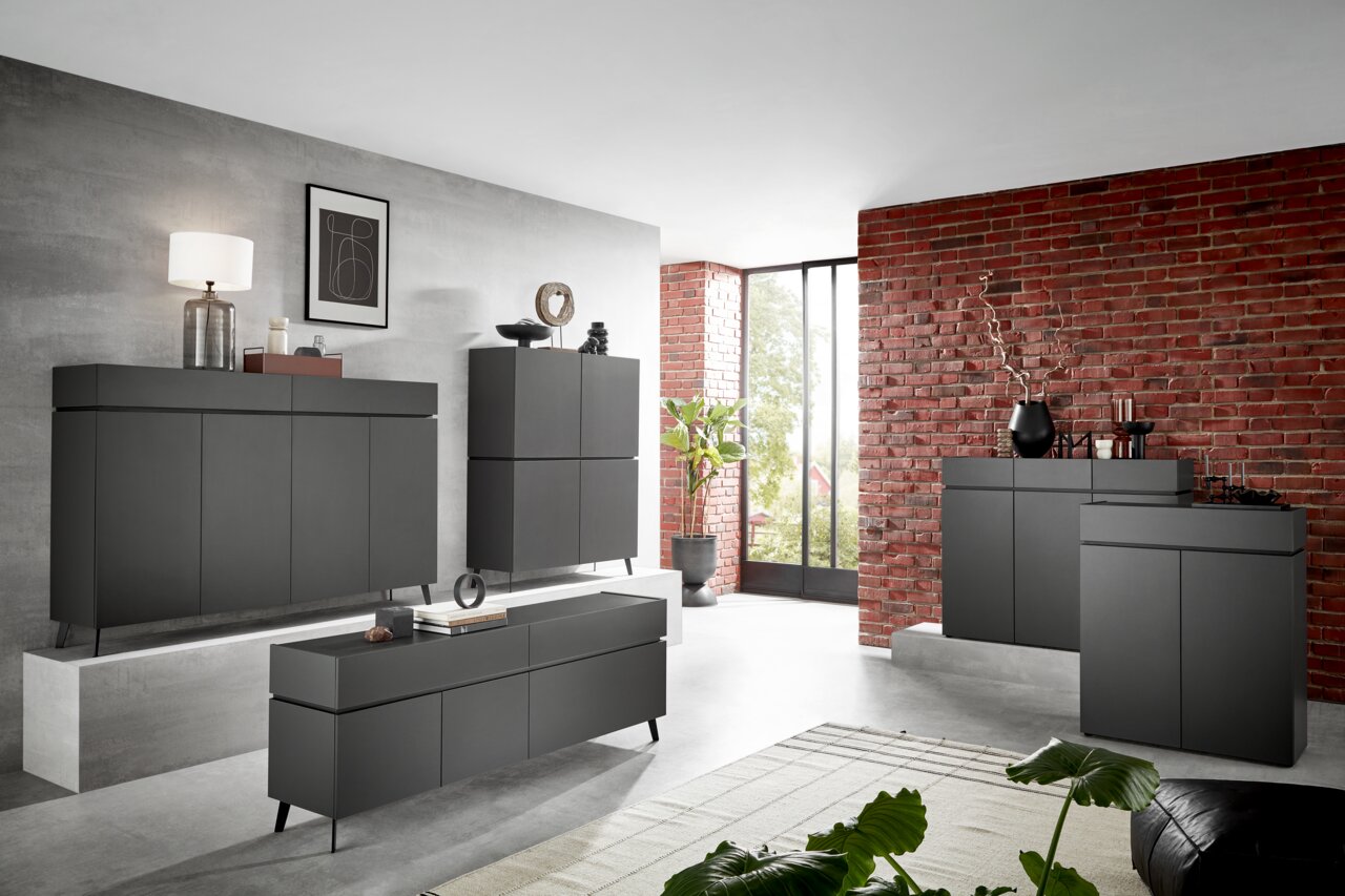 Voleo Brede ladekast UNIQUE Moderne, donkergrijze meubels in een stijlvolle woonkamer met bakstenen muur en grote ramen, gezien vanuit de zijkant.