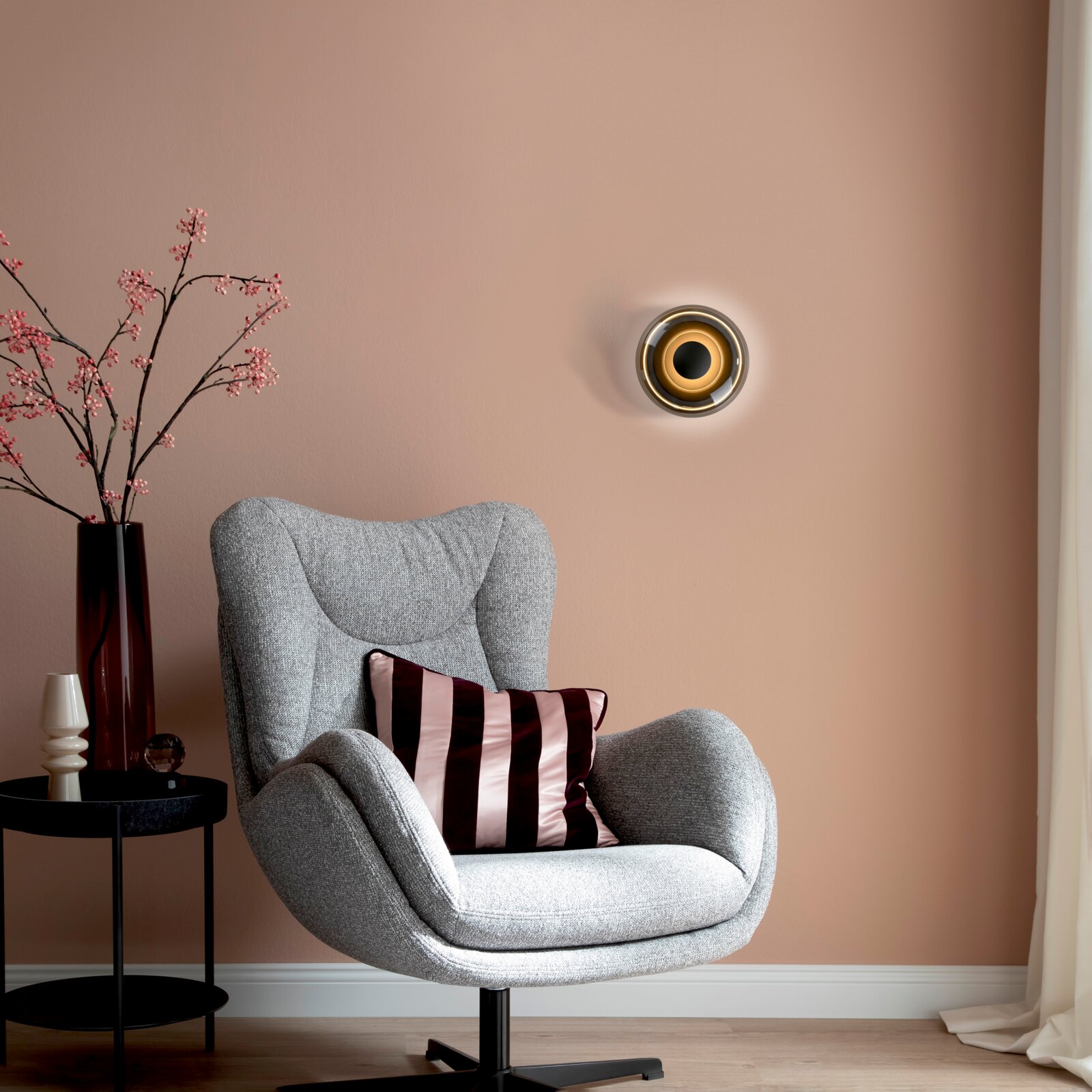 Modernes Wohnzimmer-Setting mit grauem, gepolstertem Drehsessel und gestreiftem Kissen, daneben schwarzer Beistelltisch mit Vase und Deko, an der roséfarbenen Wand eine runde Wandleuchte Perspektive: frontale Ansicht auf Augenhöhe.