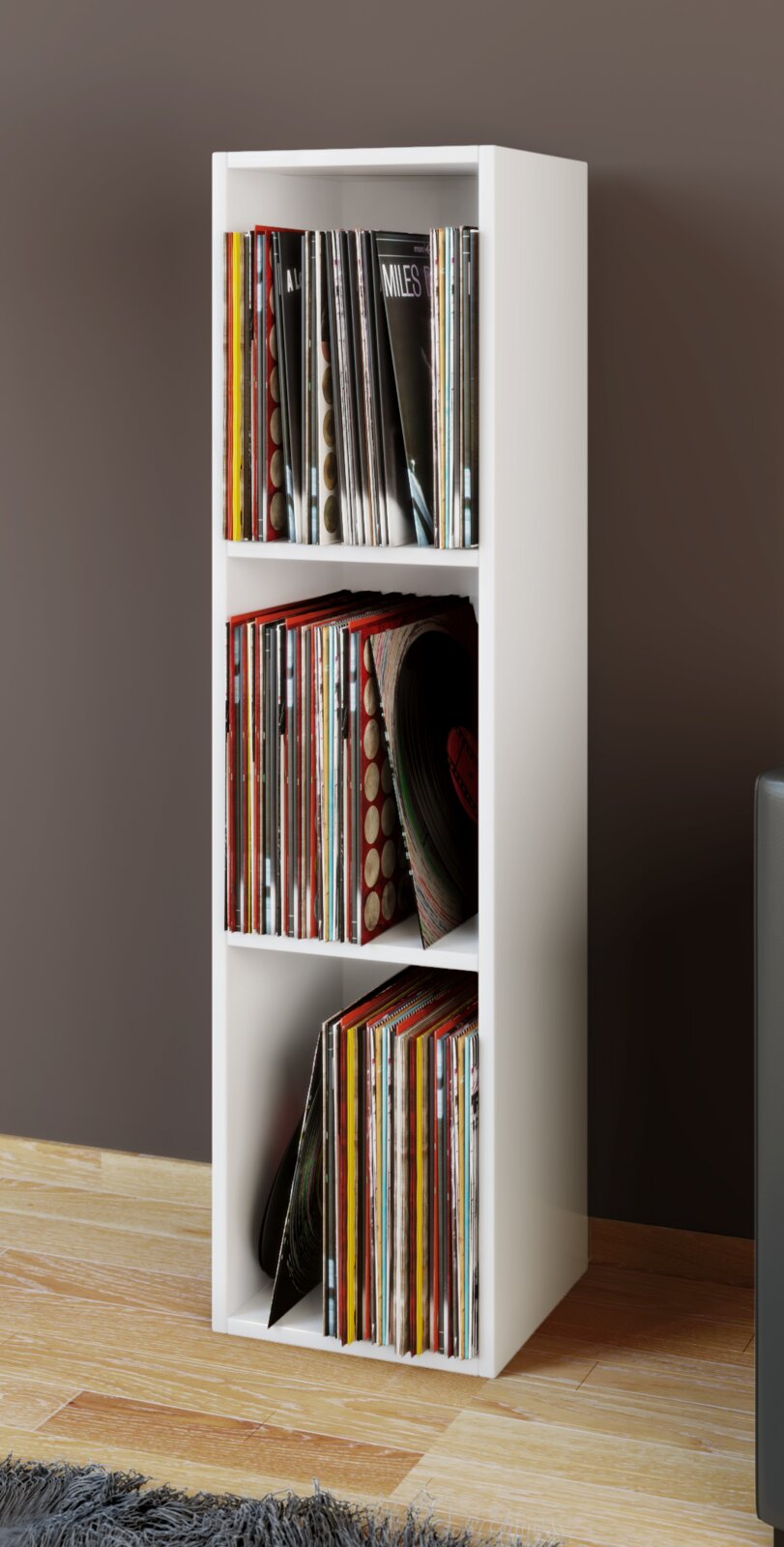 Schmales, weißes Vinyl-Schallplattenregal mit drei Fächern, gefüllt mit LPs, in schräger Frontansicht im Wohnraum auf Holzfußboden vor brauner Wand.