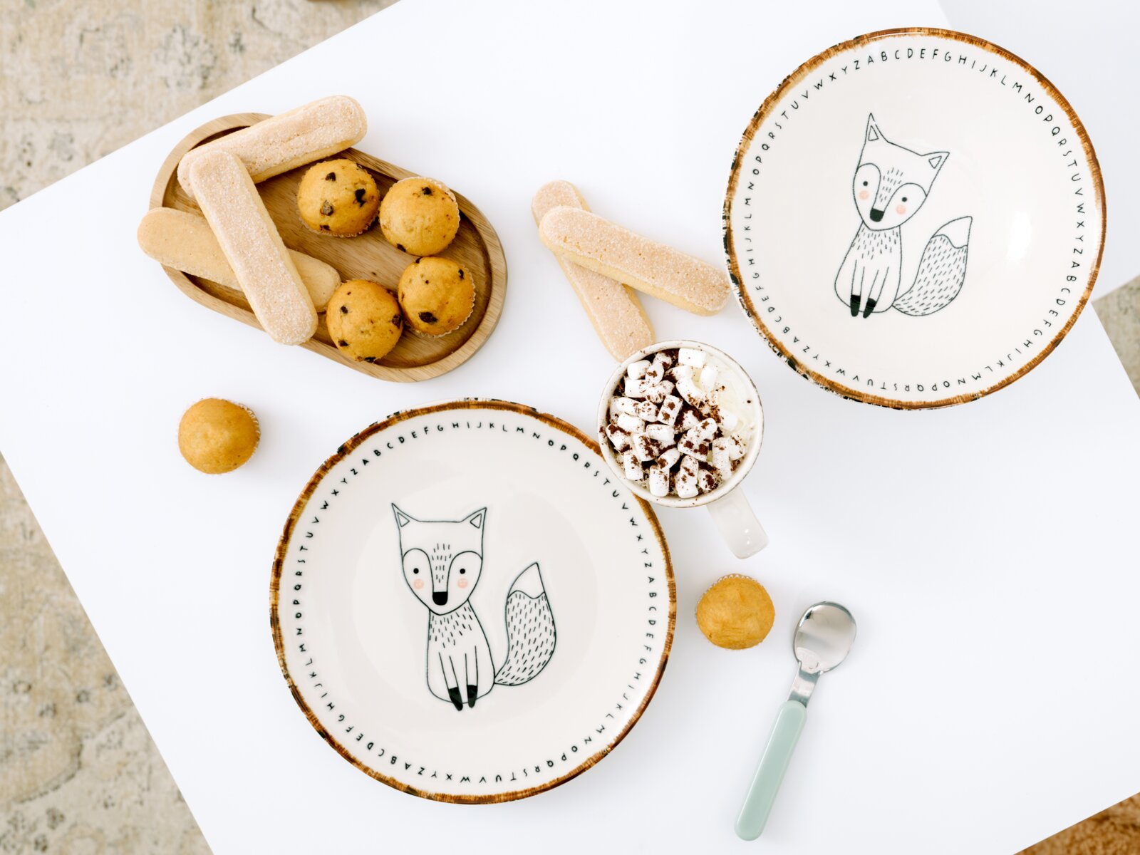 Kinder-Geschirrset mit Fuchs- und Alphabetmotiv, bestehend aus zwei Tellern und einer Tasse mit Kakao und Marshmallows, dekorativ mit Gebäck und Löffel aus der Vogelperspektive arrangiert
