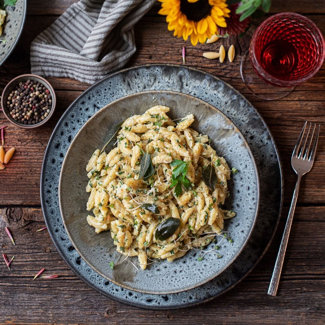 Draufsicht auf einen rustikal angerichteten Teller cremiger Spätzle-Pasta mit Kräutern und Salbei auf Holztablett, daneben Pfefferkörner, Gabel und ein Glas Rotwein