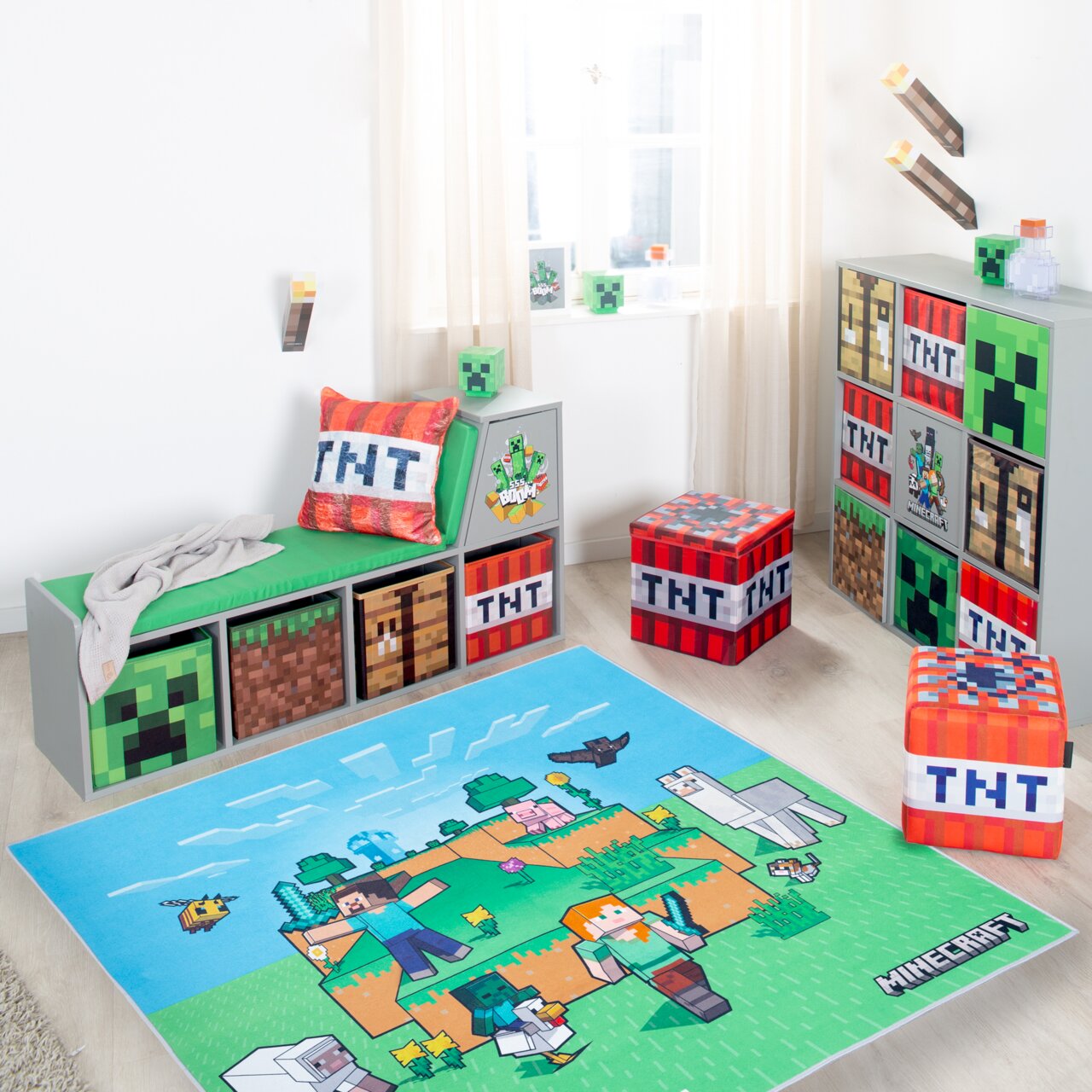 Helles Kinderzimmer im Minecraft-Design mit blau-grünem Minecraft-Teppich im Vordergrund, grauem Sitzbank-Regal mit grüner Auflage und TNT-Kissen links sowie TNT-Sitzwürfeln und Würfelregal rechts, fotografiert aus leicht erhöhter Frontperspektive.