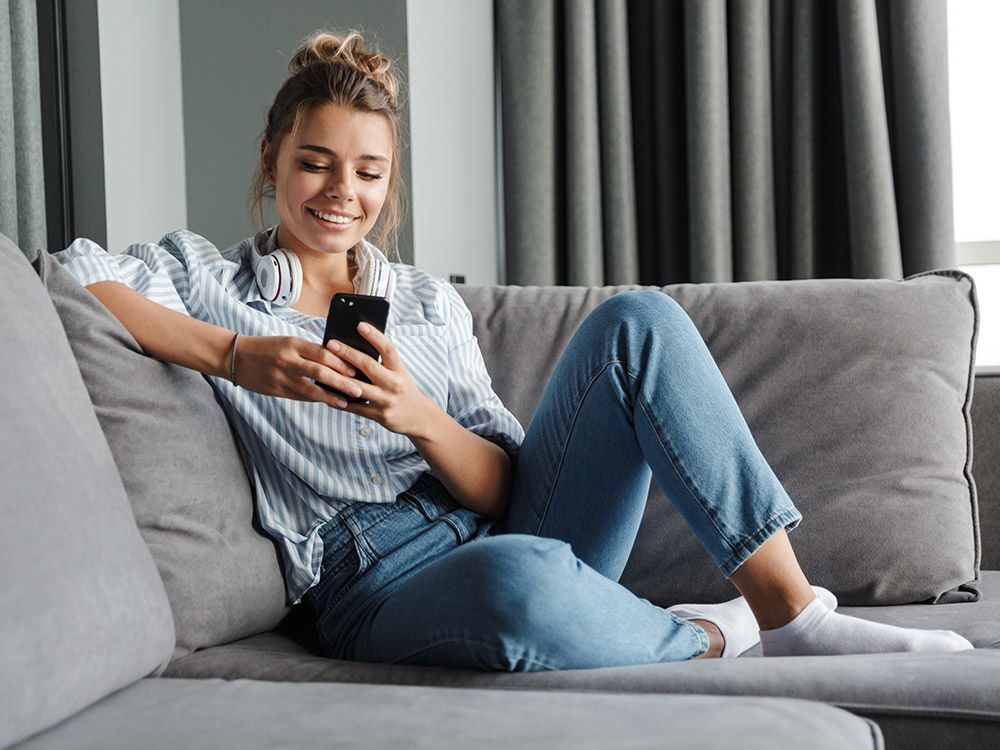 Junge Frau mit blonden Haaren sitzt entspannt auf einem grauen Sofa und schaut lächelnd auf ihr Smartphone. Sie trägt eine gestreifte Bluse, eine blaue Jeans und weiße Socken. Um den Hals hängen weiße Kopfhörer. Im Hintergrund dunkle Vorhänge und ein Fens