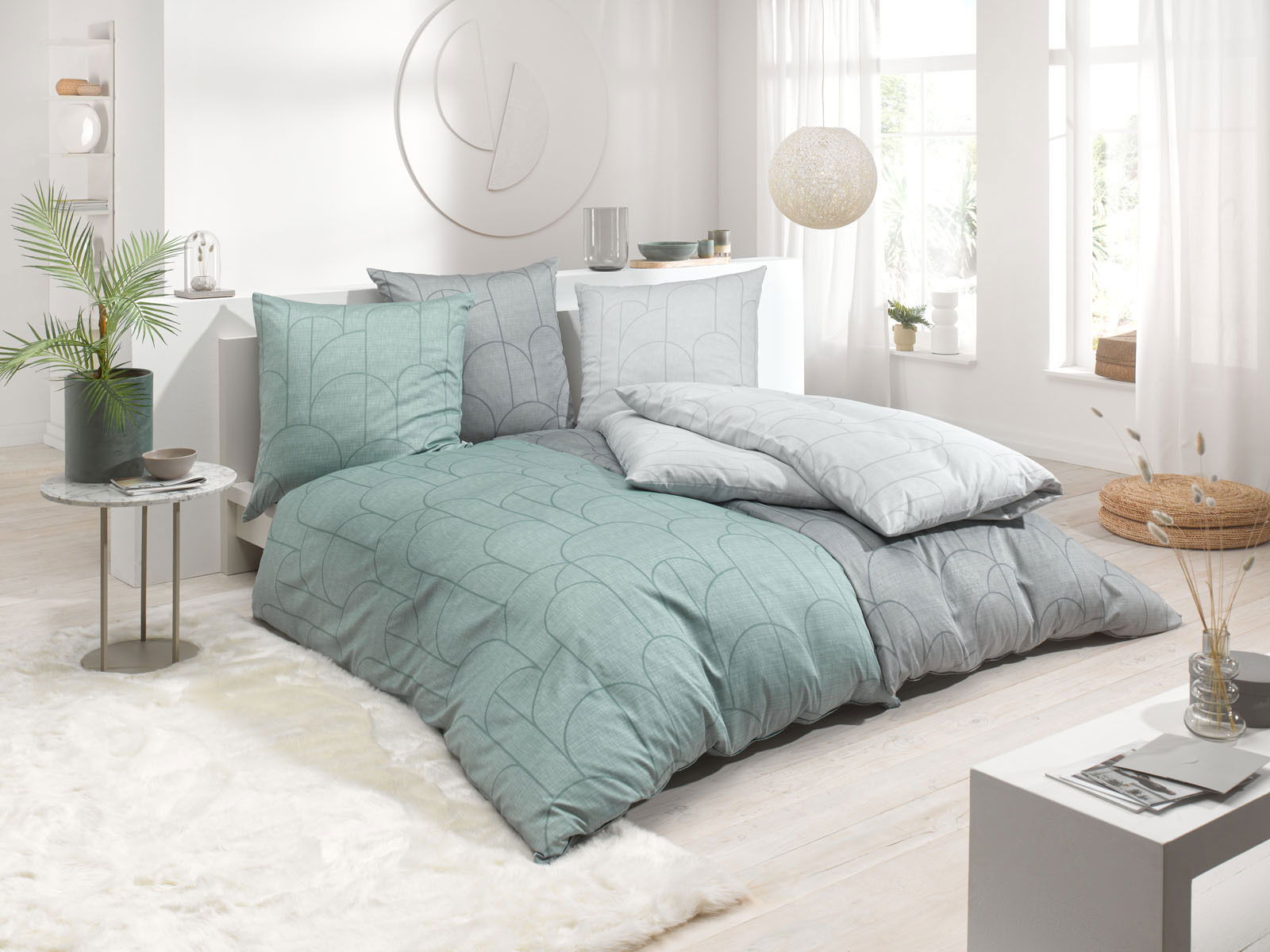 Bettwäsche-Set in einem modernen Schlafzimmer mit Tageslicht, bestehend aus Kissen und Decken in Grün- und Grautönen mit geometrischem Muster, seitliche Perspektive.