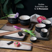 Sushi-Set für 4 Personen mit schwarzen Schalen und rechteckigen Tellern, dekoriert mit Blumen und Frühlingszwiebeln, auf einem Holztisch von oben fotografiert.