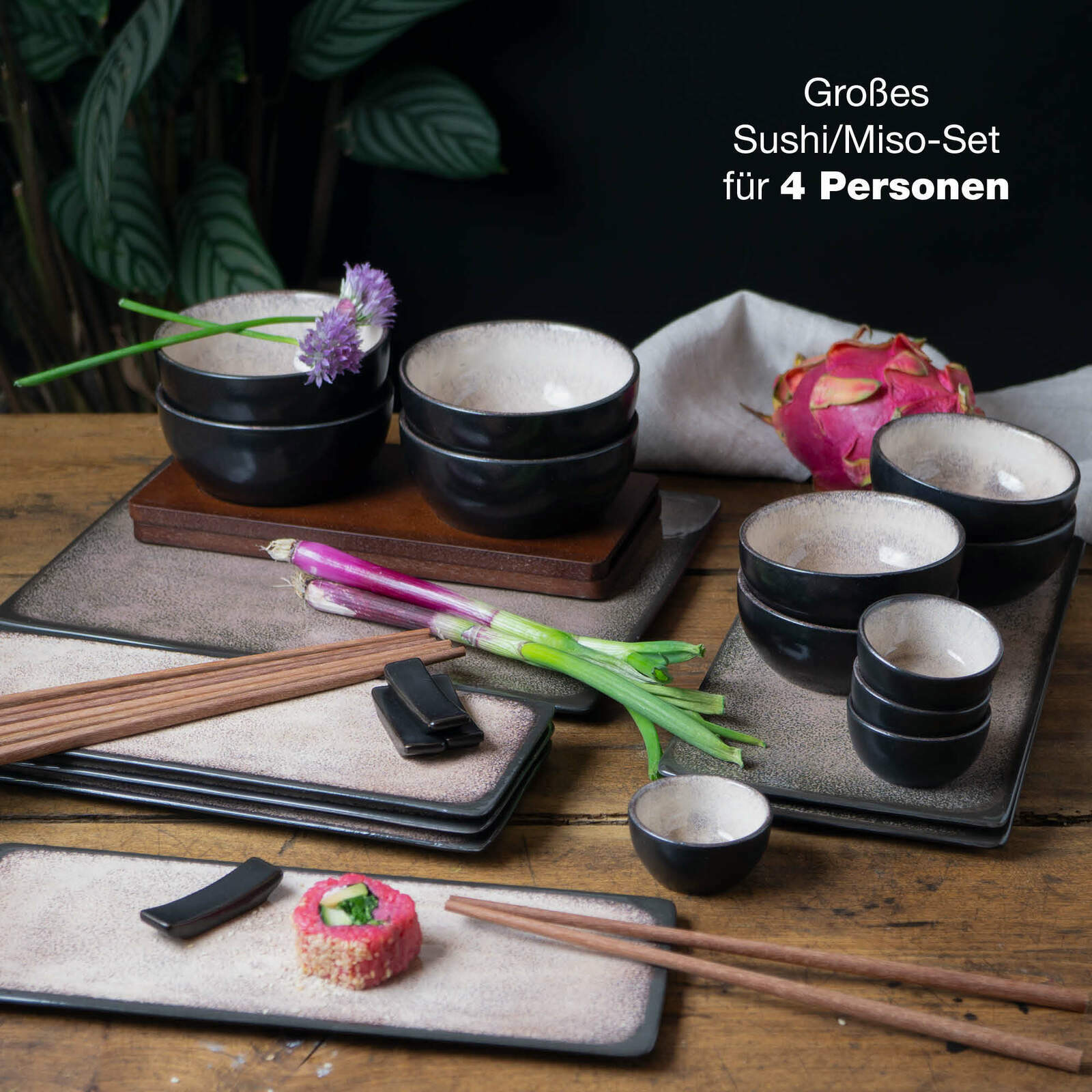 Sushi-Set für 4 Personen mit schwarzen Schalen und rechteckigen Tellern, dekoriert mit Blumen und Frühlingszwiebeln, auf einem Holztisch von oben fotografiert.