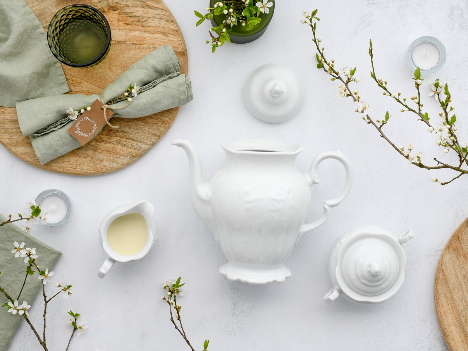 Draufsicht (Flatlay) auf ein weißes Porzellan-Teeservice mit Kanne, Zuckerdose und Milchkännchen auf hellem Untergrund, dekoriert mit Holzbrett, grünem Leinen und blühenden Zweigen