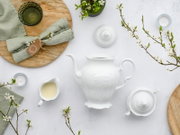 Draufsicht (Flatlay) auf ein weißes Porzellan-Teeservice mit Kanne, Zuckerdose und Milchkännchen auf hellem Untergrund, dekoriert mit Holzbrett, grünem Leinen und blühenden Zweigen