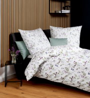 Florale Bettwäsche in Weiß mit lila und blauen Blumenmuster auf einem Bett mit dunkelgrauem Kopfteil, fotografiert in schräger Frontansicht von leicht oben; im Hintergrund Holzlamellenwand, Nachttisch mit Vase und Regal mit Büchern