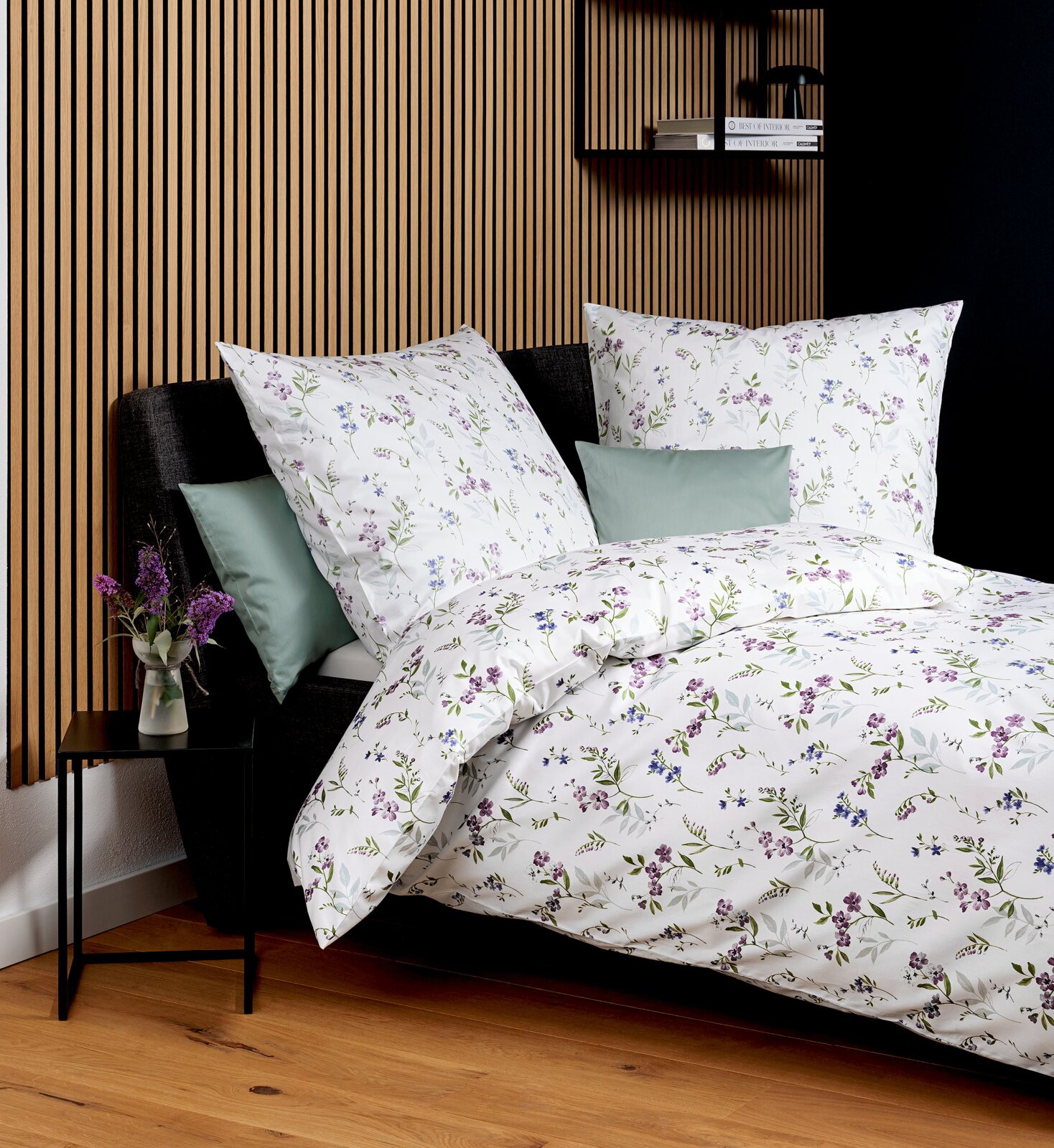 Janine Mako-Satin Bettwäsche 135/200cm PALERMO Florale Bettwäsche in Weiß mit lila und blauen Blumenmuster auf einem Bett mit dunkelgrauem Kopfteil, fotografiert in schräger Frontansicht von leicht oben; im Hintergrund Holzlamellenwand, Nachttisch mit Vase und Regal mit Büchern