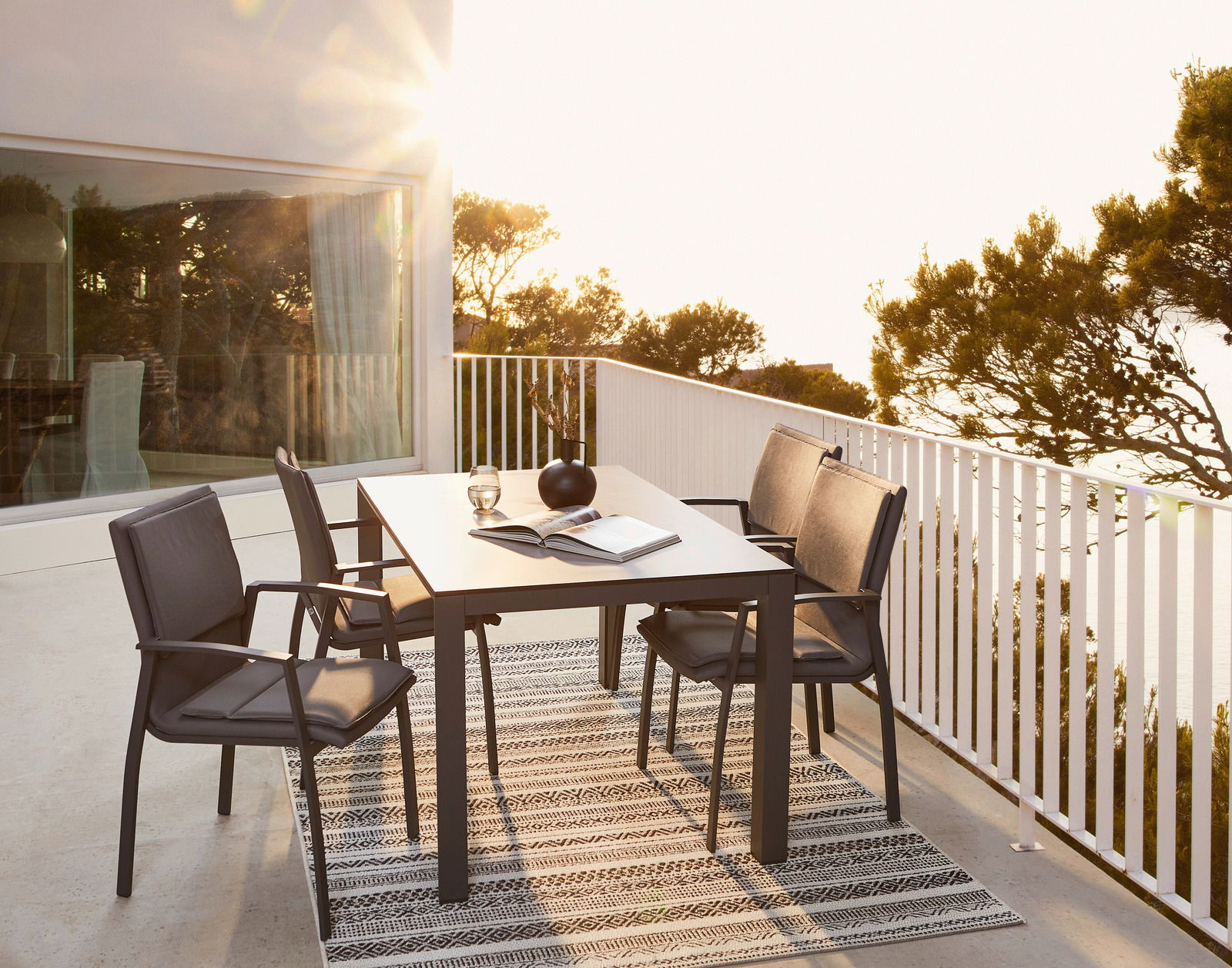 Moderner Gartentisch mit vier Stühlen auf einer Terrasse, umgeben von Bäumen und Sonnenlicht, seitliche Perspektive.