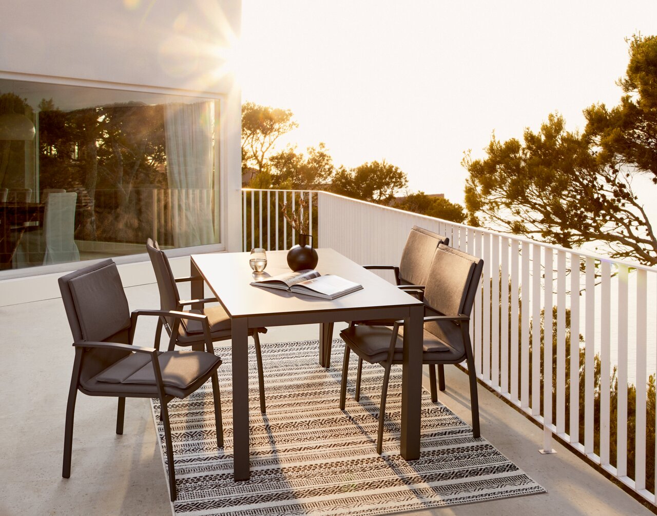 Moderne tuintafel met vier stoelen op een terras, omringd door bomen en zonlicht, zijaanzicht.