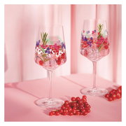 Zwei elegante Weingläser mit buntem Blumenmuster, gefüllt mit Eiswürfeln, Beeren und Rosmarin, auf einem rosa Hintergrund. Seitliche Perspektive.