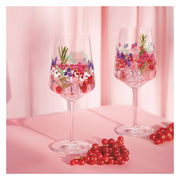 Zwei elegante Weingläser mit buntem Blumenmuster, gefüllt mit Eiswürfeln, Beeren und Rosmarin, auf einem rosa Hintergrund. Seitliche Perspektive.