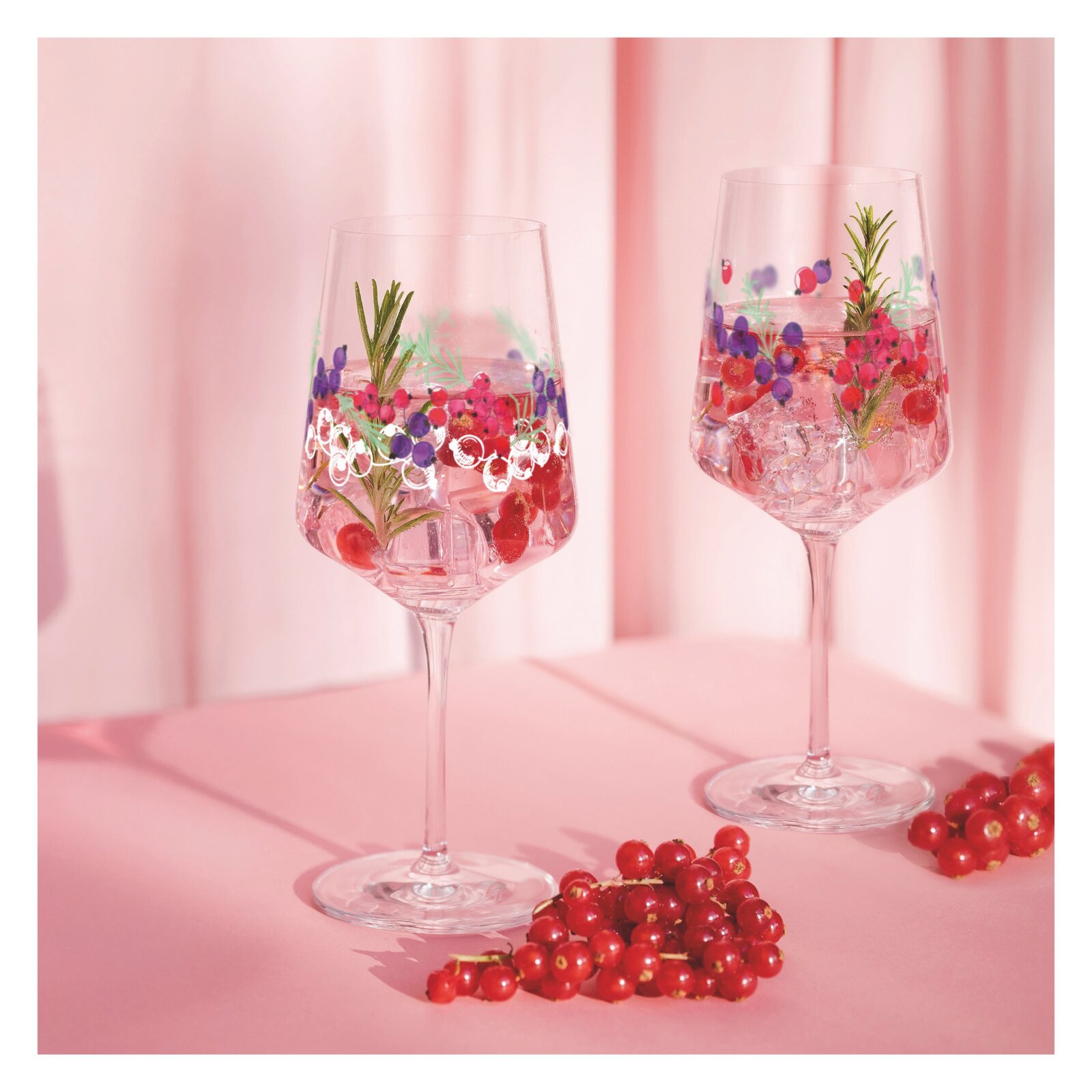 Zwei elegante Weingläser mit buntem Blumenmuster, gefüllt mit Eiswürfeln, Beeren und Rosmarin, auf einem rosa Hintergrund. Seitliche Perspektive.